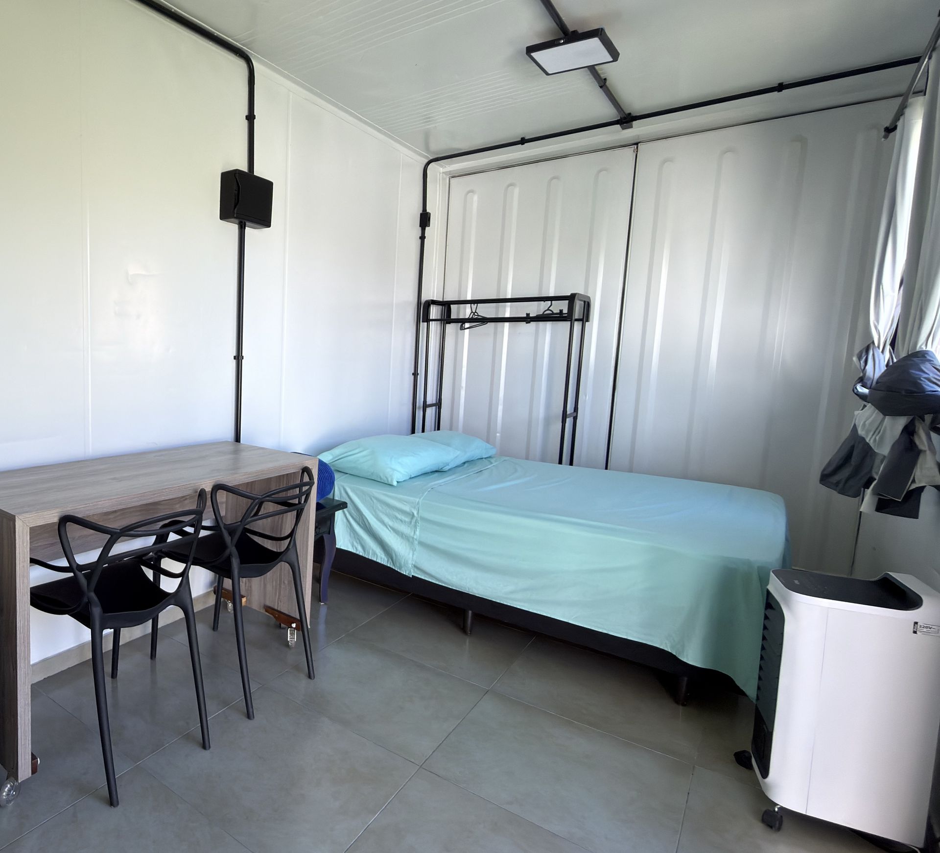 Suíte do container com a cama feita em lençóis verde-água, mesa com duas cadeiras e arara para roupas.