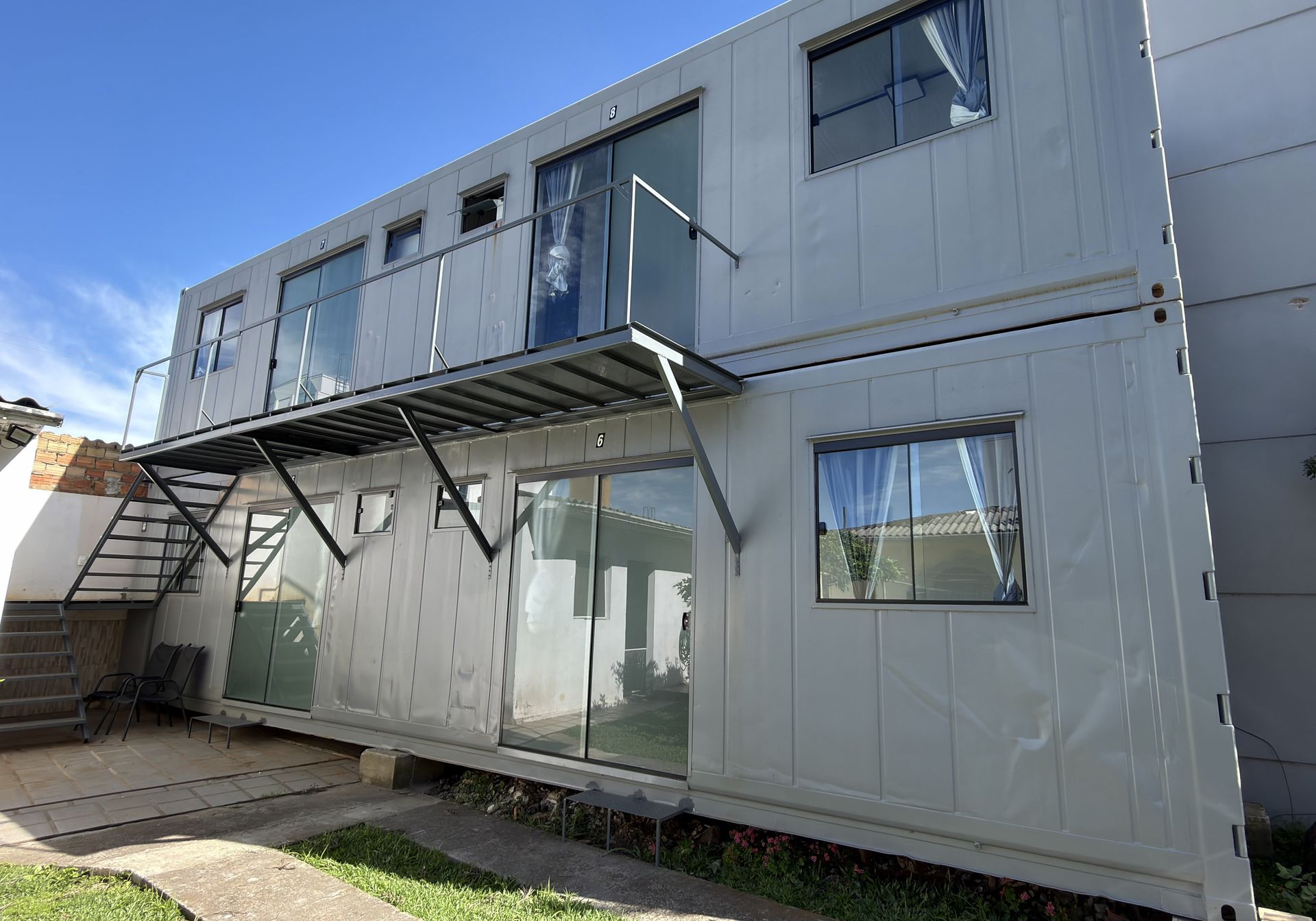 As suítes em container, em dois andares, com sacada, portas de vidro e o gramado na frente.