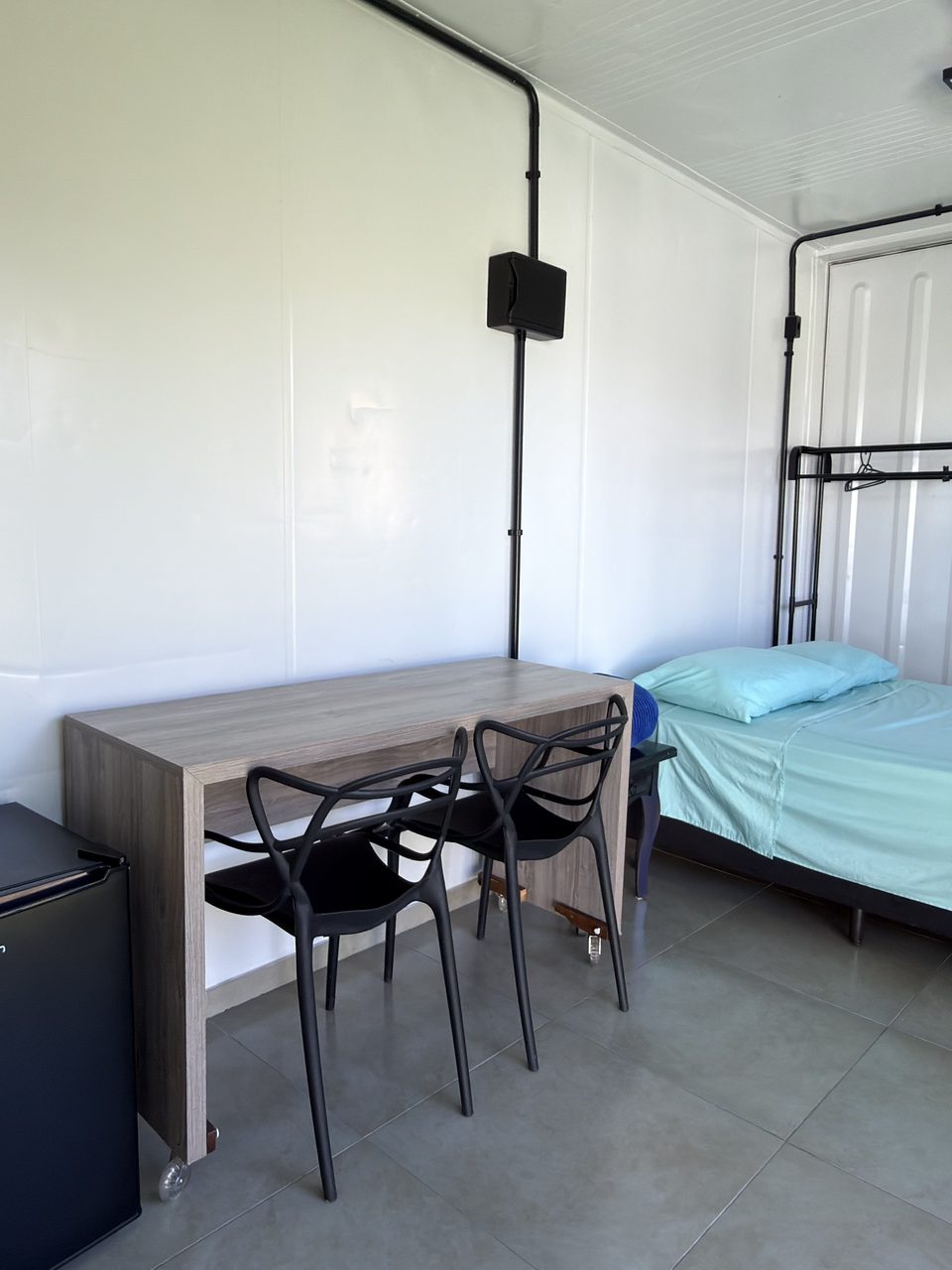 O Quarto 6 por dentro: a cama com roupa verde-água, a mesa de trabalho e o frigobar.